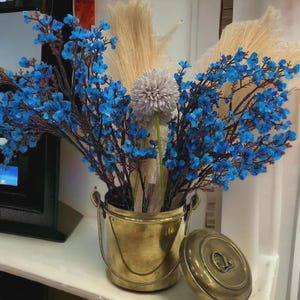 Puede incluir: Un cubo de latón con tapa, lleno de flores azules artificiales y hierbas beige secas. Una sola flor gris también está incluida en el arreglo. El cubo está sobre una superficie blanca.