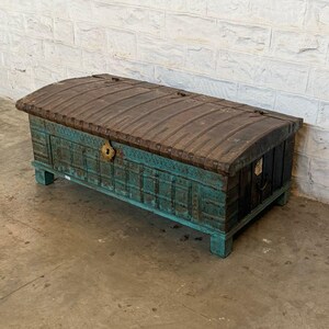 May include: A large, antique wooden chest with a curved, dark brown top and teal-colored sides. The chest features intricate carvings and a brass-colored lock. The chest is rectangular and sits on short, square feet.