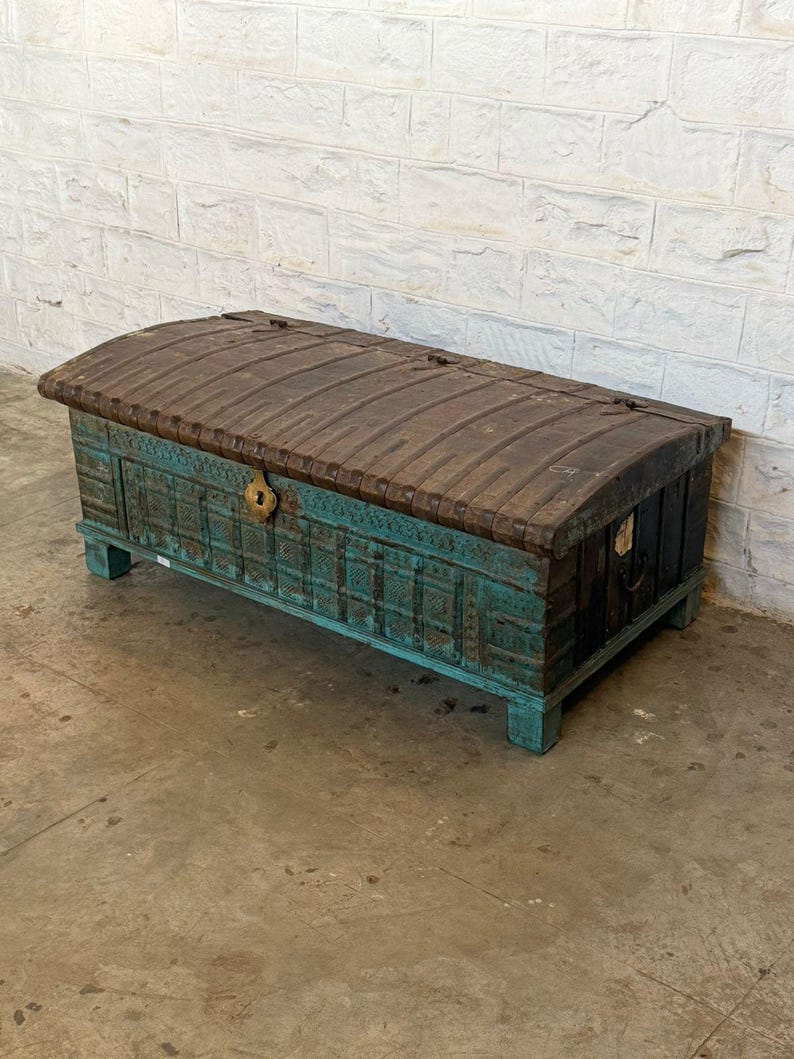 May include: A large, antique wooden chest with a curved, dark brown top and teal-colored sides. The chest features intricate carvings and a brass-colored lock. The chest is rectangular and sits on short, square feet.