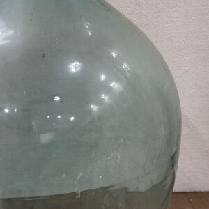 May include: A large, clear glass bottle with a rounded body and a narrow neck. The bottle has a slightly greenish tint and appears to be vintage. The glass surface reflects light, creating highlights and shadows. The bottle is likely used for decorative purposes.