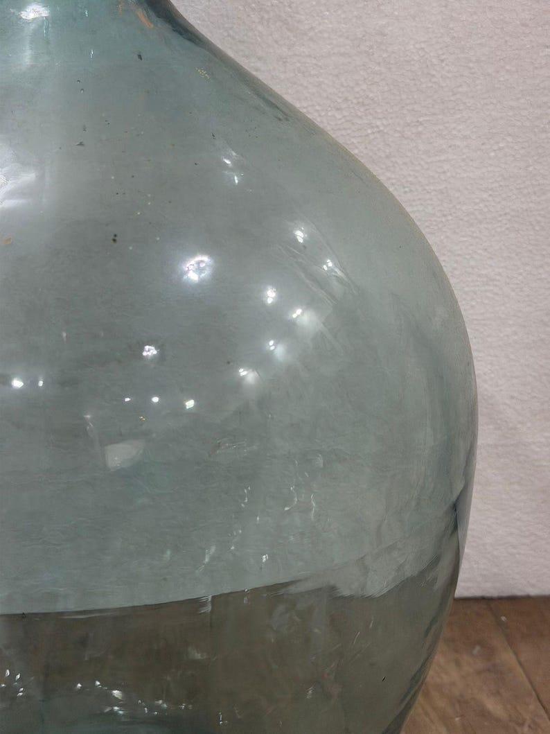 May include: A large, clear glass bottle with a rounded body and a narrow neck. The bottle has a slightly greenish tint and appears to be vintage. The glass surface reflects light, creating highlights and shadows. The bottle is likely used for decorative purposes.