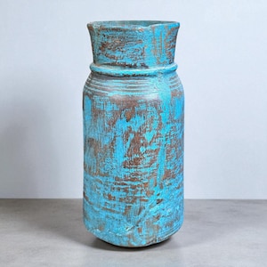 Puede incluir: Un jarrón alto y rústico con un acabado turquesa desgastado. El jarrón tiene un cuerpo cilíndrico con una parte superior ligeramente ensanchada y una banda alrededor del cuello. La veta de la madera es visible a través de la pintura, dándole un aspecto vintage. Este jarrón decorativo es adecuado para la decoración del hogar.