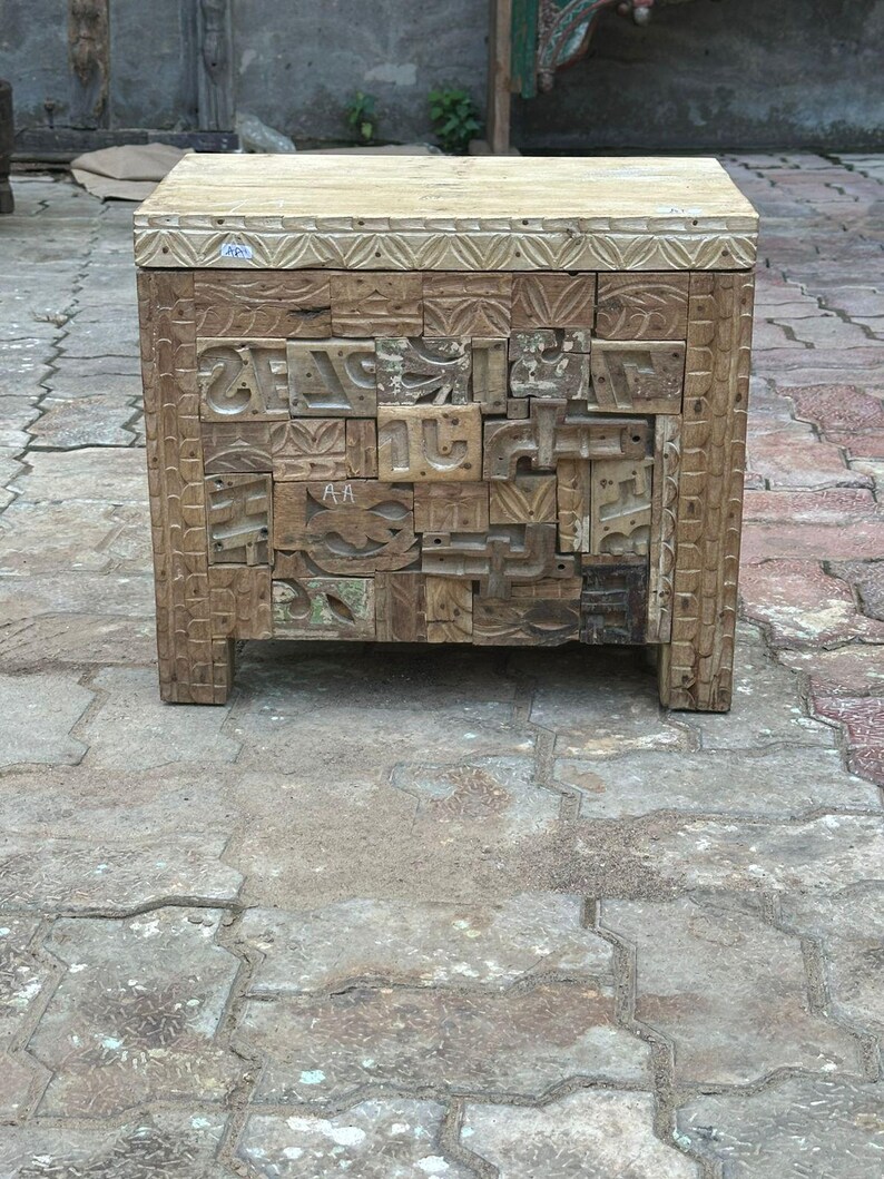 Op de afbeelding: Een handgemaakte houten bijzettafel met een lichtgekleurd blad en een ingewikkeld gesneden voorkant. De voorkant bestaat uit verschillende houten blokken met gesneden patronen en letters. De tafel heeft een natuurlijke, verweerde uitstraling.