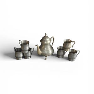 Op de afbeelding: Een vintage zilveren theeservies, bestaande uit een theepot en verschillende kleine kopjes. De theepot heeft een decoratief bloemmotief en een gebogen tuit. De kopjes hebben kleine handvatten. De set wordt gepresenteerd tegen een witte achtergrond.