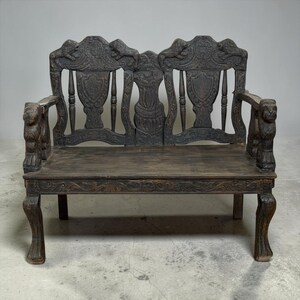 May include: A dark brown wooden bench featuring intricate carvings of lions on the armrests and top rail. The backrest includes two shield-shaped panels and a central carved pillar. The bench has four legs with carved details and a flat seat.