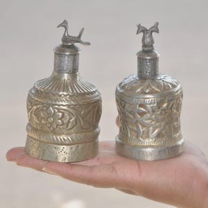 May include: Two antique silver-colored bottles with ornate floral and bird designs. The bottles have detailed carvings and decorative stoppers. The bottles are held in a person's hand.