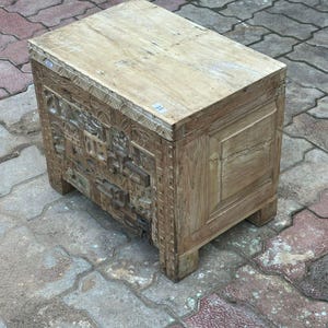 Op de afbeelding: Een rechthoekige houten kist met een lichtbruine afwerking. De voorkant van de kist is voorzien van gedetailleerde houtsnijwerken, terwijl de bovenkant een glad, vlak oppervlak is. Hij staat op korte, vierkante poten en heeft een vintage of antiek ontwerp. De kist is ongeveer 60 cm lang.