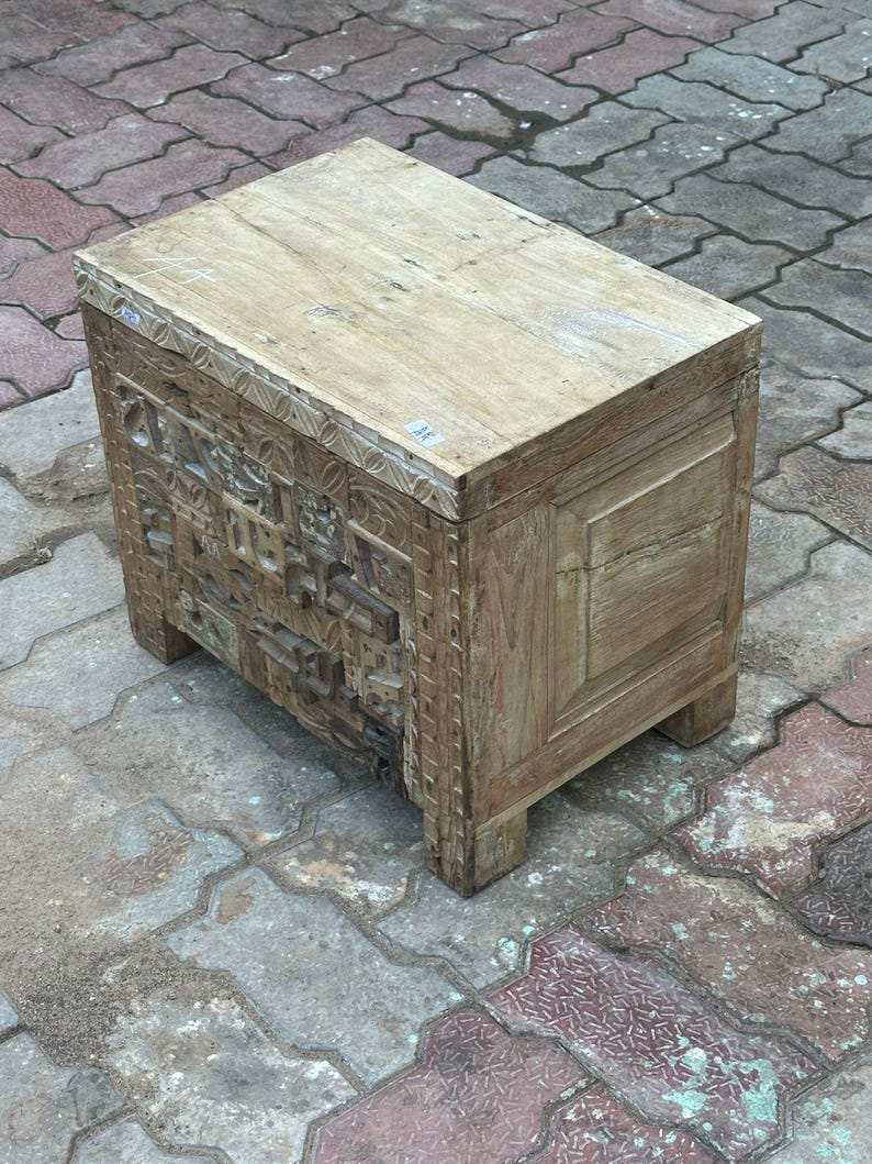Op de afbeelding: Een rechthoekige houten kist met een lichtbruine afwerking. De voorkant van de kist is voorzien van gedetailleerde houtsnijwerken, terwijl de bovenkant een glad, vlak oppervlak is. Hij staat op korte, vierkante poten en heeft een vintage of antiek ontwerp. De kist is ongeveer 60 cm lang.