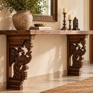 May include: A wooden console table featuring a thick, rectangular top and two ornate, carved supports. Each support displays detailed relief carvings of birds and foliage. The table surface holds a large textured vase, books, and candles.