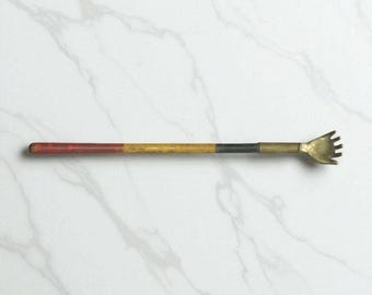 May include: A back scratcher with a wooden handle, painted red, yellow, and black. The scratching end is a small, hand-shaped piece of metal. The handle is approximately 35.5 cm long.