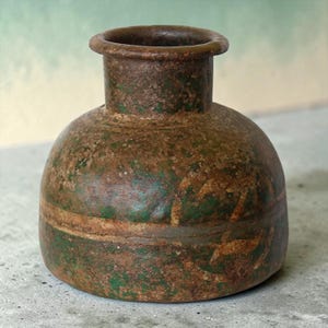 May include: A small, round, wide-mouthed vessel with a textured, weathered surface. The exterior features a mottled appearance with patches of deep brown, rust-like tones and vibrant green pigment, showing a horizontal seam around the center.