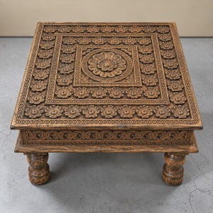 May include: A square wooden coffee table featuring intricate hand-carved floral patterns across the top surface and side panels. The table is finished in a medium brown stain and is supported by four thick, rounded, carved wooden legs.