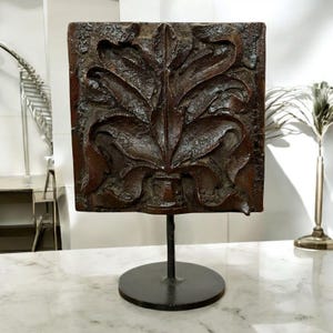 May include: A dark brown, square, carved wooden panel with a stylized floral design. It is mounted on a black metal stand with a circular base. The detailed carving has a textured appearance.