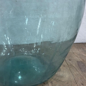 May include: A large, clear glass vase with a wide, rounded shape. The top portion has a textured, frosted appearance, transitioning to a transparent, teal-colored base. The vase sits on a wooden surface.