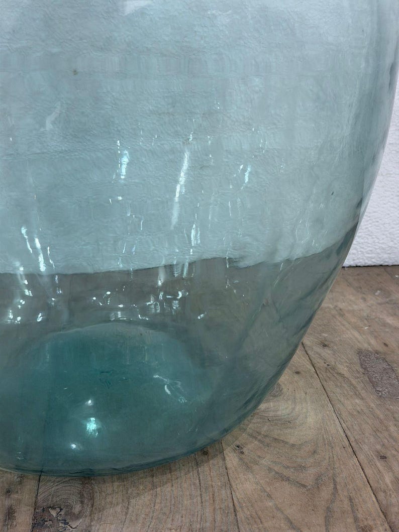May include: A large, clear glass vase with a wide, rounded shape. The top portion has a textured, frosted appearance, transitioning to a transparent, teal-colored base. The vase sits on a wooden surface.