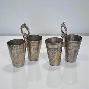 May include: Four antique metal cups with decorative etched patterns. Each cup is silver-toned with a gold-colored band and a handle with a bird-like design. The cups are arranged in pairs.