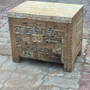 Op de afbeelding: Een verweerde houten bijzettafel met ingewikkelde gesneden details. De tafel heeft een rechthoekige vorm met een lichtgekleurd bovenblad en een voorpaneel met een moza&iuml;ek van gesneden blokken. Hij wordt ondersteund door vier korte poten.