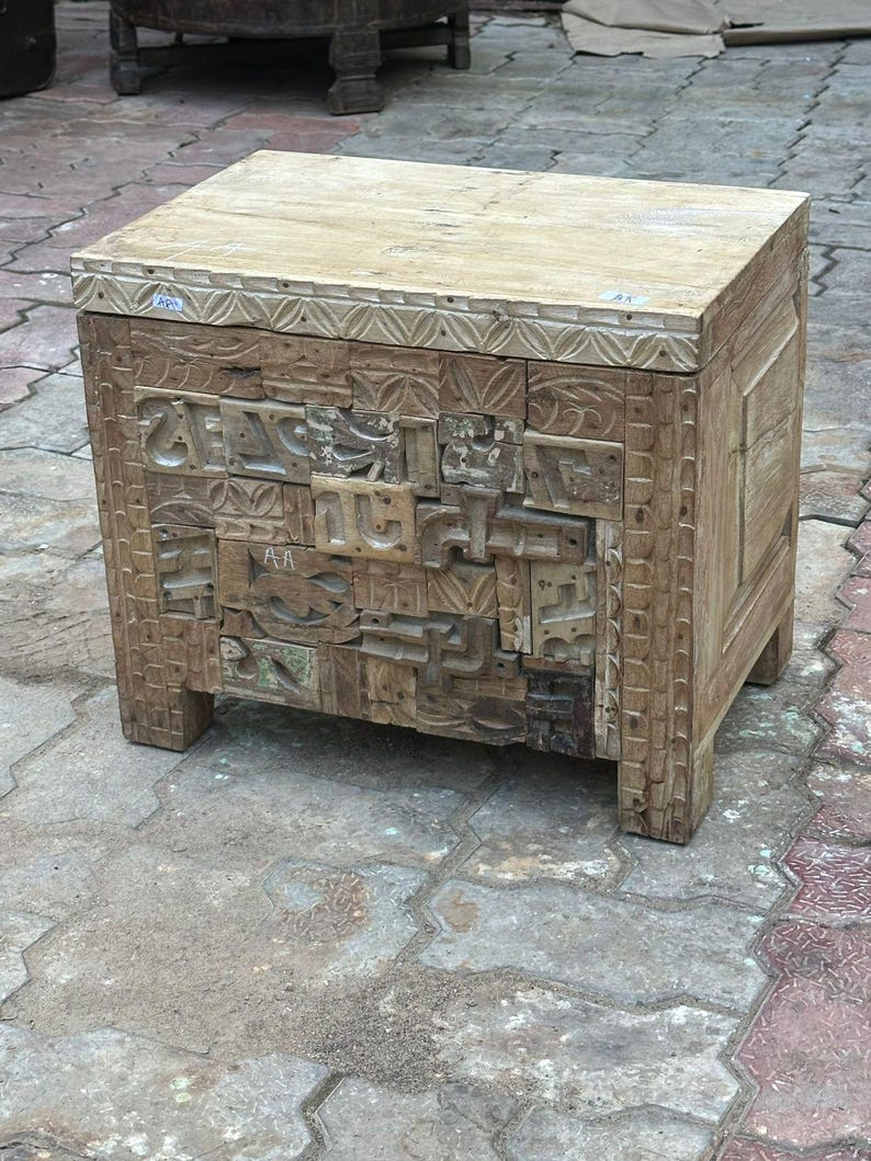Op de afbeelding: Een verweerde houten bijzettafel met ingewikkelde gesneden details. De tafel heeft een rechthoekige vorm met een lichtgekleurd bovenblad en een voorpaneel met een moza&iuml;ek van gesneden blokken. Hij wordt ondersteund door vier korte poten.