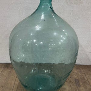 May include: A large, clear glass demijohn with a wide, rounded body and a narrow neck. The glass has a slight teal tint and is partially frosted, creating a two-tone effect. The demijohn sits on a wooden surface.