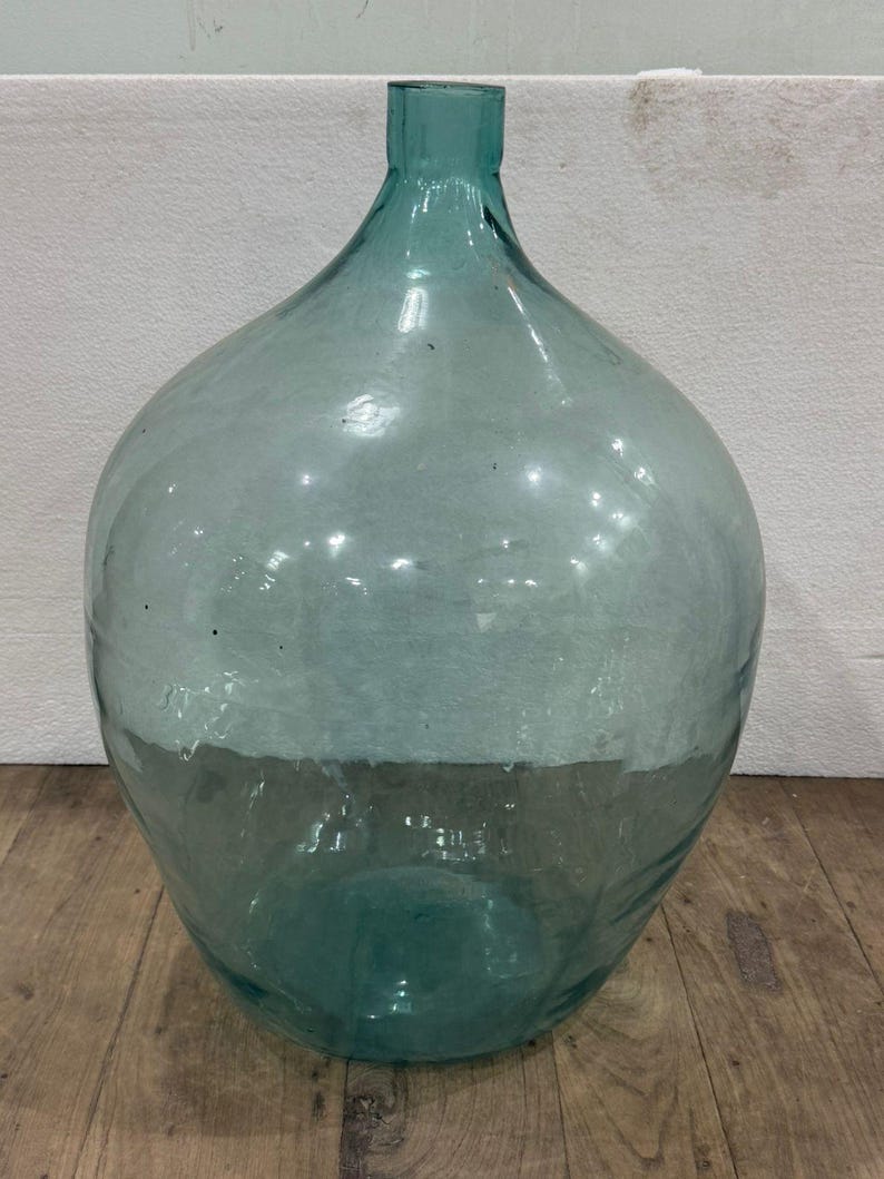 May include: A large, clear glass demijohn with a wide, rounded body and a narrow neck. The glass has a slight teal tint and is partially frosted, creating a two-tone effect. The demijohn sits on a wooden surface.