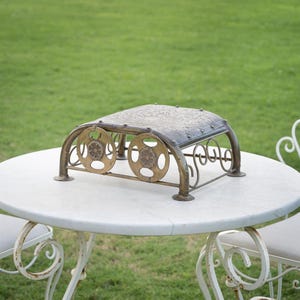 May include: An antique-style metal footstool with a rectangular top and decorative floral patterns. The footstool has a bronze finish with two circular side accents and curved supports. It is placed on a white marble table with a green grass background.