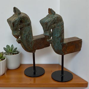 Könnte beinhalten: Zwei antike Holzskulpturen von Pferdeköpfen mit einer verwitterten, türkis-braunen Oberfläche. Jeder Pferdekopf ist auf einem schwarzen Metallständer mit rundem Sockel montiert. Die Skulpturen werden auf einem Holzregal ausgestellt.