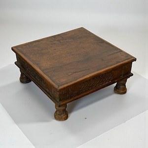 May include: A square, wooden footstool with a dark brown finish. The top surface is flat and the edges are slightly raised. The stool rests on four short, rounded legs. The sides feature carved decorative elements.