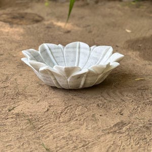 May include: A white marble bowl shaped like a flower, with petal-like edges. The bowl is sitting on a sandy surface. The bowl is a decorative item.
