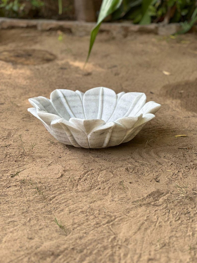May include: A white marble bowl shaped like a flower, with petal-like edges. The bowl is sitting on a sandy surface. The bowl is a decorative item.