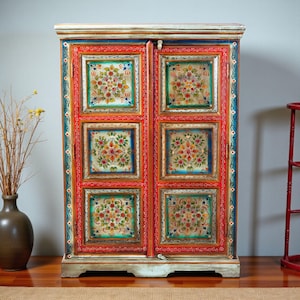 May include: A vibrantly painted wooden cabinet with two doors. The doors feature floral designs within square panels, framed by red, blue, and green borders. The cabinet's base and top are painted in a light, distressed finish, and the overall style is reminiscent of folk art.