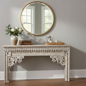 May include: A white, intricately carved wooden console table with a decorative border and floral accents. A gold-framed round mirror hangs above the table. The table displays a vase of white flowers, books, and a tea set.