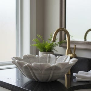 May include: A marble flower-shaped bathroom sink with a gold faucet. The sink is light gray with darker gray veining. The faucet is curved and has a lever handle. A small potted plant sits behind the sink.