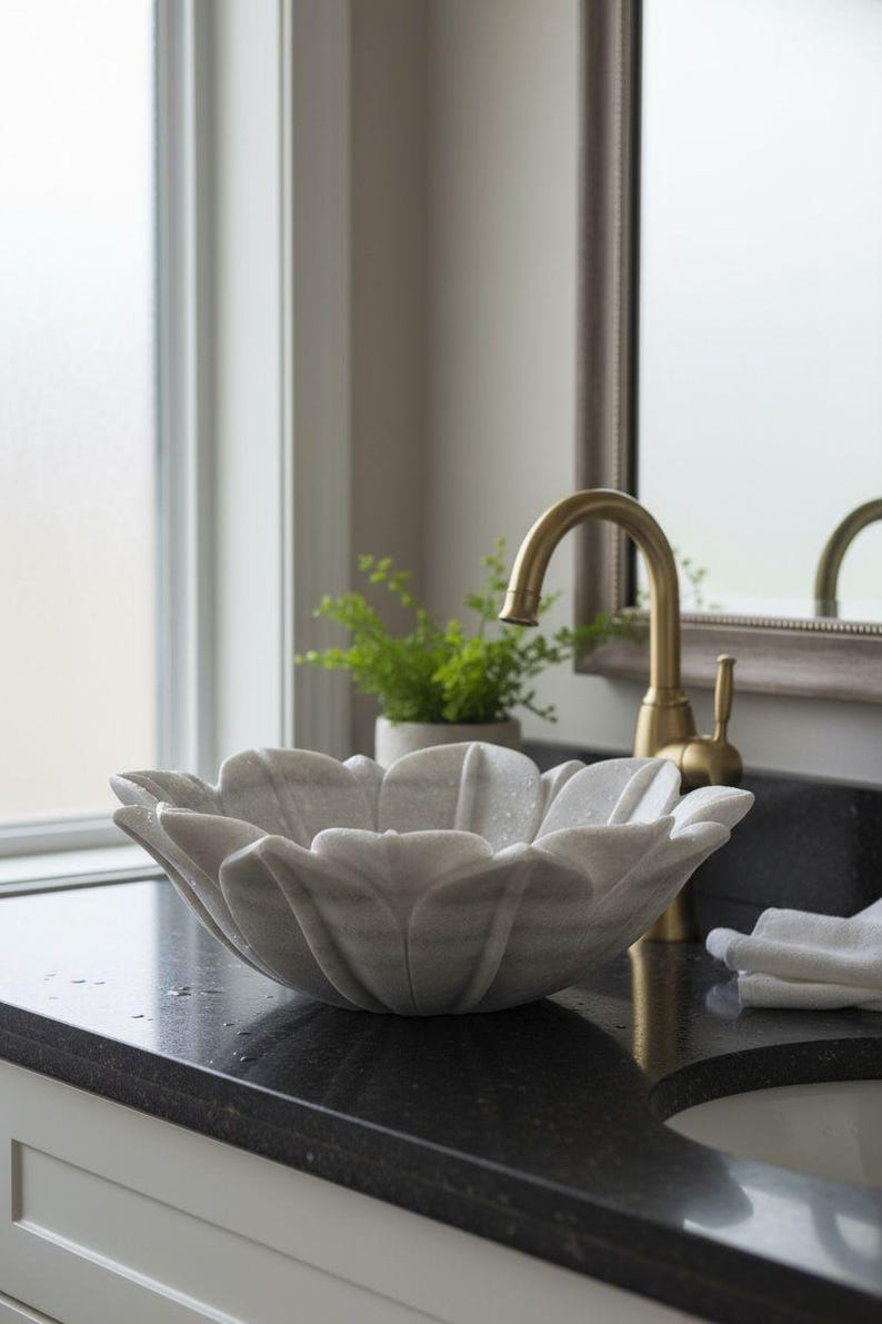 May include: A marble flower-shaped bathroom sink with a gold faucet. The sink is light gray with darker gray veining. The faucet is curved and has a lever handle. A small potted plant sits behind the sink.