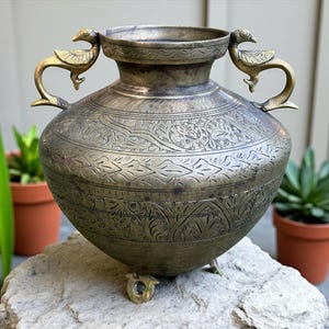 May include: Vintage brass vase with detailed etched floral designs. The vase has two bird-shaped handles and three small feet. The aged metal has a dark, tarnished look, enhancing its vintage appeal. A decorative item for home decor.