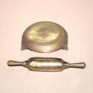 May include: A polished brass rolling pin with textured handles and a matching round brass tray featuring an ornate floral engraved border. The tray is supported by three small feet, and both items exhibit a metallic gold finish with visible patina.
