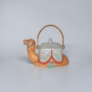 May include: A ceramic teapot shaped like a camel, with a tan body and a white, tent-like structure on its back. The teapot has a woven handle and is decorated with red, green, and blue accents. The camel's head is detailed with a blue bridle.