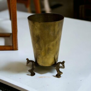 May include: A vintage brass vase with a flared top and a decorative base. The vase has a warm, golden hue and a slightly aged appearance. The base features three ornate, curved legs.