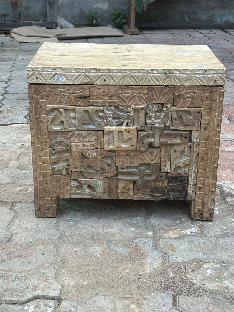 Op de afbeelding: Een handgemaakte houten bijzettafel met een lichtgekleurd blad en ingewikkeld gesneden zijkanten. De body van de tafel is samengesteld uit diverse houten blokken, elk met unieke patronen en texturen. De tafel wordt ondersteund door vier stevige poten.