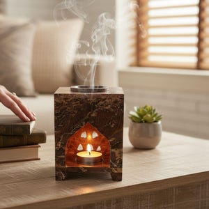 Peut inclure: Un diffuseur d'huiles essentielles en marbre brun avec une bougie allumée à l'intérieur. Le diffuseur a une fenêtre arquée décorative et un dessus en métal. De la fumée s'élève du dessus. Une petite plante succulente en pot et des livres sont à proximité.