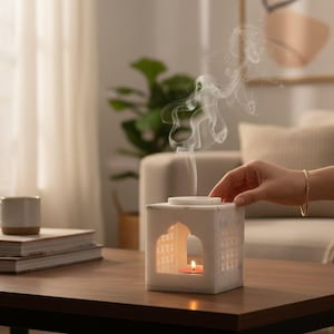 Könnte beinhalten: Ein weißer, würfelförmiger Diffusor für ätherische Öle mit einem Gebäude-Silhouetten-Design. Im Inneren brennt eine kleine Kerze, und Dampf steigt auf. Der Diffusor steht auf einem dunkelbraunen Holztisch.