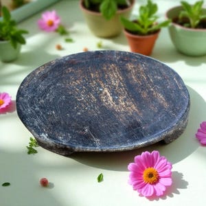 May include: A dark, oval-shaped wooden cutting board with a distressed finish. The board has a dark brown and gray color with visible wood grain. Pink flowers and small potted plants surround the board, suggesting a rustic or natural theme.