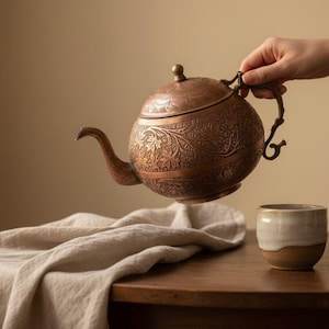 May include: A copper-colored teapot with intricate floral patterns is held above a small, two-toned ceramic cup. The teapot has a curved spout and a decorative handle. A beige cloth rests on a wooden table.