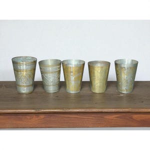 May include: Five vintage metal drinking glasses with a silver and gold finish. The glasses are arranged in a row on a wooden surface. Each glass has a unique etched design, adding to their antique charm.