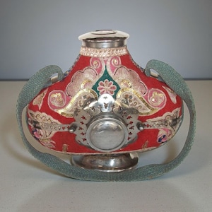 May include: A decorative red fabric flask with silver detailing and a matching lid. The flask features gold and pink embroidery, sequins, and a green strap. The base and central element are silver.