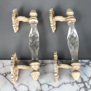 Puede incluir: Dos manijas de puerta antiguas con un centro de vidrio transparente y facetado y una ornamentada metalistería color crema. Las manijas tienen un diseño clásico, perfecto para agregar un toque de elegancia vintage a cualquier puerta. Sobre una superficie de mármol.