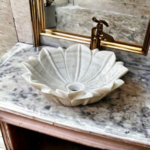 May include: A white marble sink carved in a flower shape. The sink is set on a marble countertop with a gold faucet and mirror frame in the background. The sink has a central drain hole.