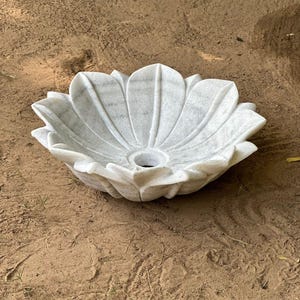 May include: A white marble lotus flower-shaped sink. The sink has a central drain hole and is made of white marble with gray veining. The sink is designed to resemble a blooming lotus flower, with detailed petal edges.