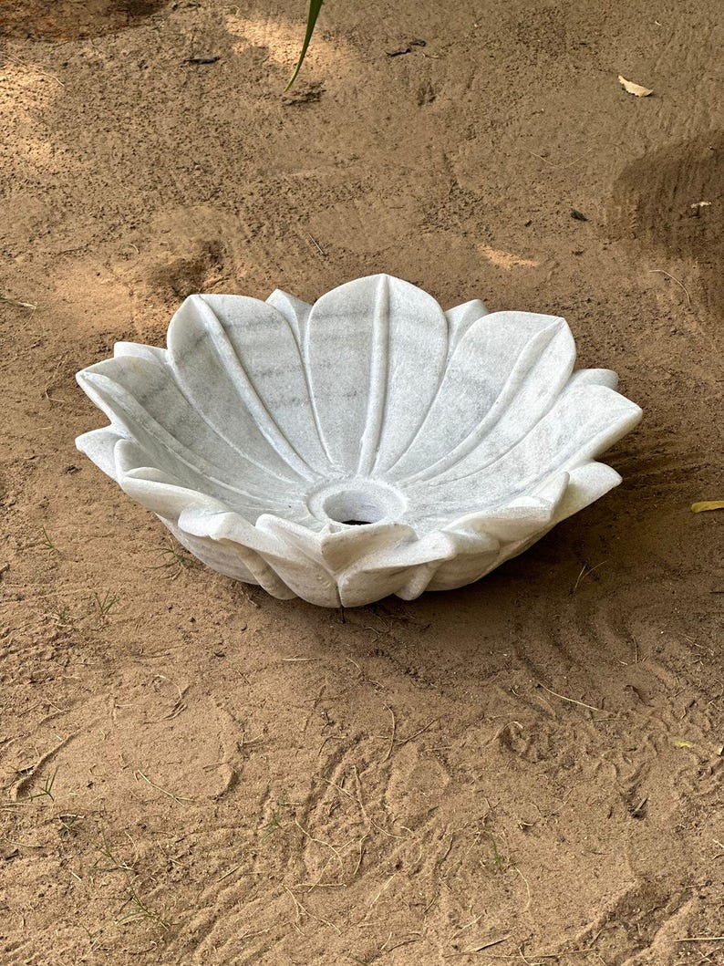 May include: A white marble lotus flower-shaped sink. The sink has a central drain hole and is made of white marble with gray veining. The sink is designed to resemble a blooming lotus flower, with detailed petal edges.
