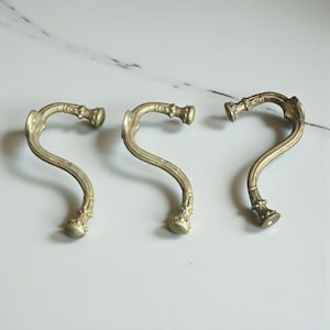 Pode incluir: Três ganchos de parede antigos, cor de latão, com um design curvo e ornamentado. Cada gancho tem um adorno decorativo na parte superior e inferior, e um corpo curvo em forma de S. Os ganchos estão dispostos sobre uma superfície de mármore branco.