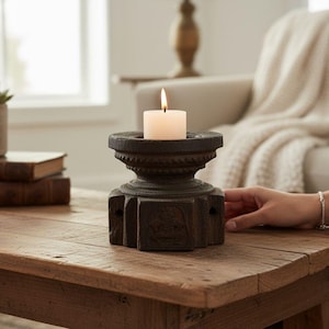 Może przedstawiać: Płonąca biała świeca w ciemnobrązowym, rzeźbionym drewnianym świeczniku. Świecznik stoi na rustykalnym drewnianym stole. Na stole stoi również sukulent w doniczce i książki.